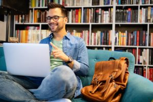 Junger Mann it Laptop auf seinem ofa, im Hintergrund Buchregale