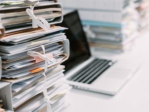 Hoher Stapel von Aktenmappen, dahinter ist unscharf ein Laptop zu erkennen.
