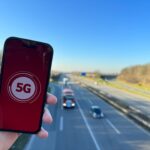 Blick von einer Autobahnbrücke, im Vordergrund eine Hand, die ein Handy mit 5G-Logo hochhält.