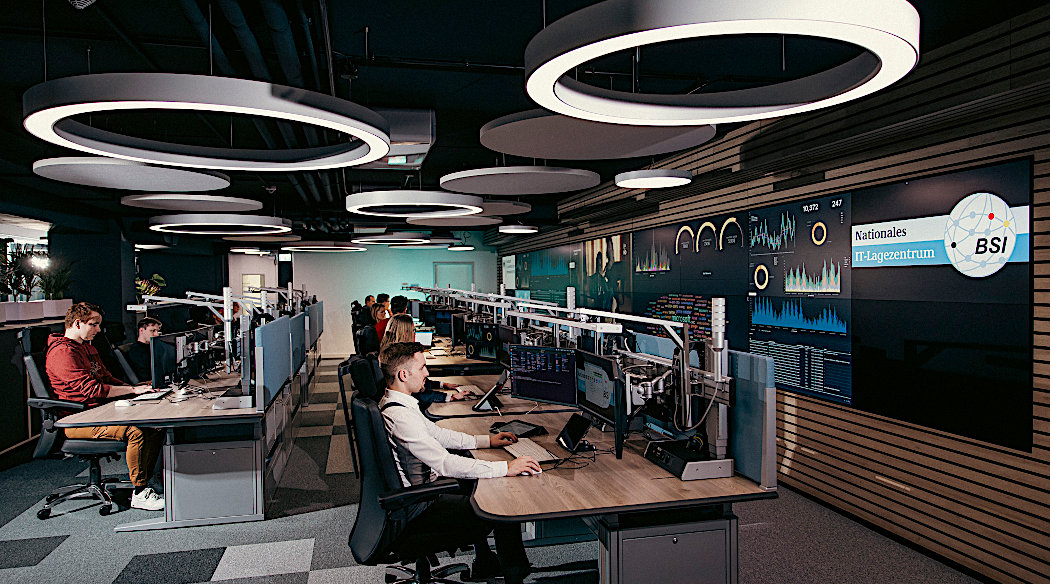 Das Bild zeigt das Innere des IT-Lagezentrums mit Personen an Bildschirmen und großen Monitoren an den Wänden.
