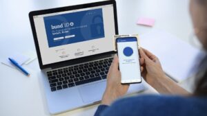 Das Bild zeigt eine Person mit Smartphone in der Hand vor einem Laptop mit geöffneter Website zur BundID.