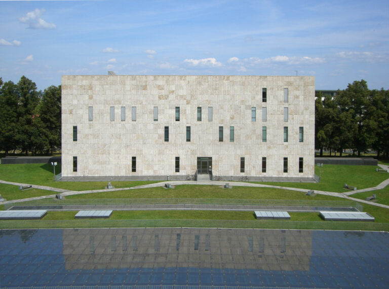 Außenansicht der SLUB, ein quadratisches, mehrstöckiges Flachdachgebäude mit ein paar, unregelmäßig angebrachten quadratischen Fenstern. Davor Wiese und der Anschnitt eines flachen Brunnens, der ebenfalls als Rechteck angelegt ist.