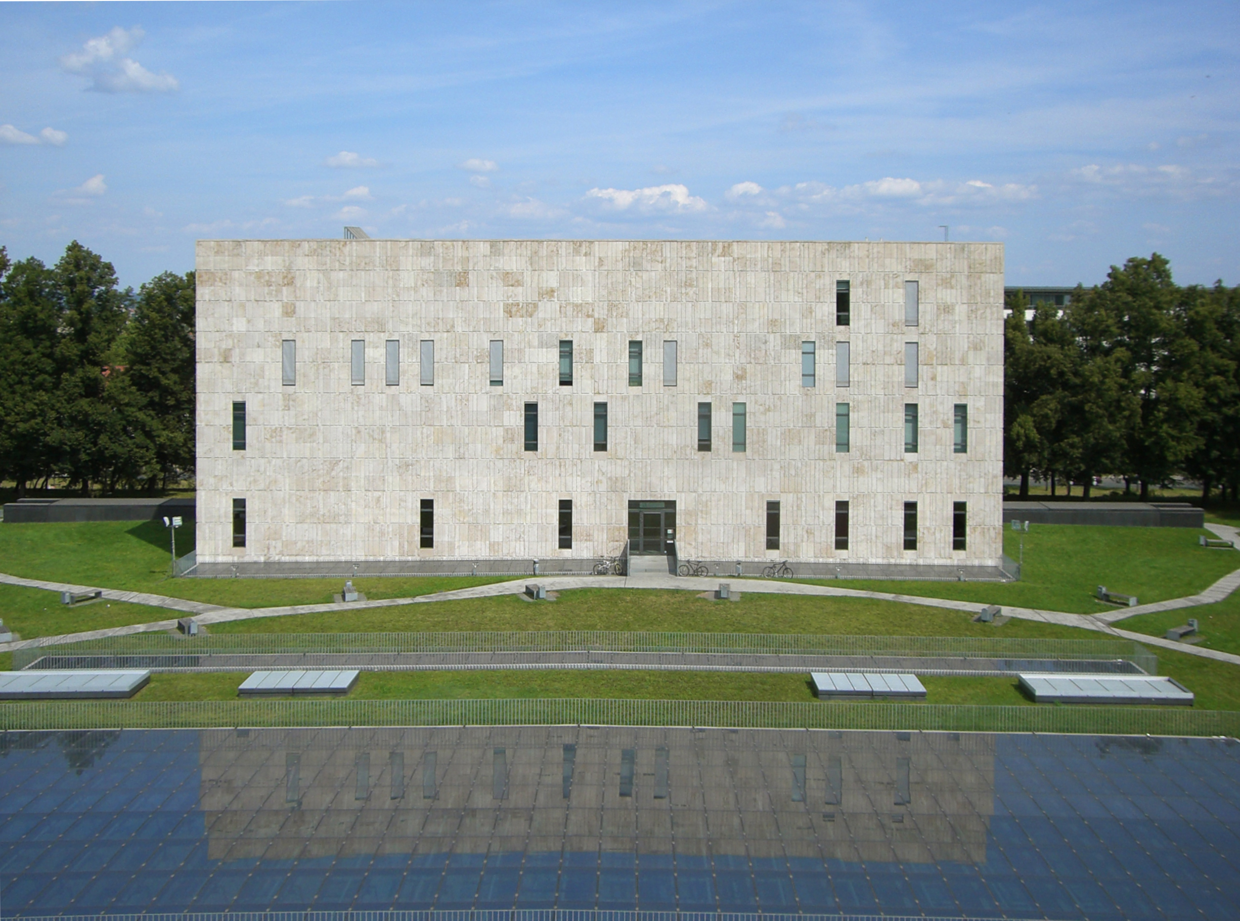 Außenansicht der SLUB, ein quadratisches, mehrstöckiges Flachdachgebäude mit ein paar, unregelmäßig angebrachten quadratischen Fenstern. Davor Wiese und der Anschnitt eines flachen Brunnens, der ebenfalls als Rechteck angelegt ist.