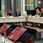 Konferenzsituation mit U-förmigem Tisch, im Vordergrund zwei Monitore, weiter hinten Menschen in Business-Kleidung.