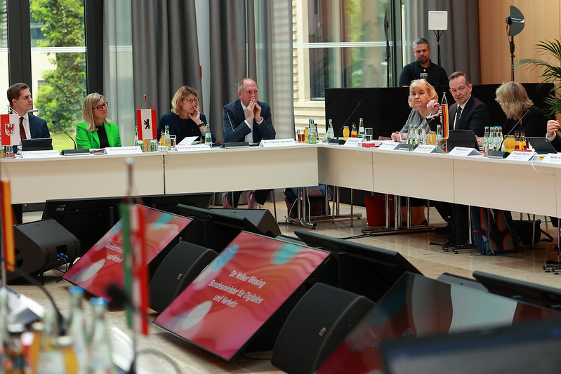 Konferenzsituation mit U-förmigem Tisch, im Vordergrund zwei Monitore, weiter hinten Menschen in Business-Kleidung.