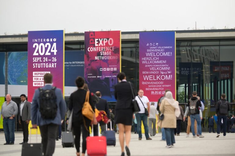 Außenansicht Messehalle mit purpurfarbenen Werbebannern für die Intergeo 2024. Eine Anzahl von Besuchern läuft auf die Eingänge zu.