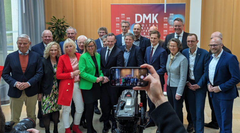 Gruppenbild der DMK-Teilnehmenden. Man sieht etwa 20 Personen in Business-Kleidung zusammenstehen, im Hintergrund Holzvertäfelung und DMK-Banner.