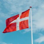 Eine rote Flagge mit weißem Kreuz (Dänemark-Flagge) weht vor blauem Himmel von rechts nach links.