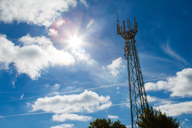 Mobilfunkmast vor blauem, leicht bewölktem Himmel im Gegenlicht.