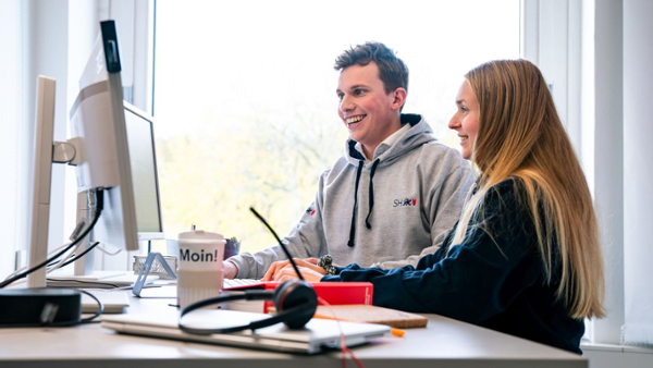 Zwei junge Menschen (männlich und weiblich) sitzen gemeinsam in einem Büro vor einem Computer-Bildschirm.