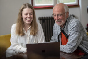 Eine junge Frau und ein älterer, grauhaariger Mann sitzen gemeinsam vor einem Laptop, auf dessen Bildschirm sie blicken.