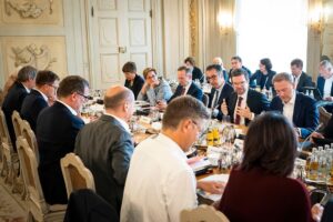 Historischer Saal mit heller, freundlicher Ausgestaltung; zahlreiche förmlich gekleidete Personen sitzen debattierend an langen Tischen.