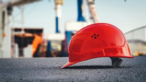 Ein roter Bauhelm liegt auf dem Asphaltboden, im Hintergrund unscharf eine Baustelle.