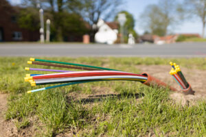 Nahaufnahme einer Wiese, im Hintergrund eine Straße. Glasfaserkabel ragen aus der Erde.