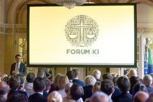Blick über Köpfe und Schulter enes Publikums hinweg auf eine Vortragsbühne, dort ist das Logo "Forum KI" projiziert.