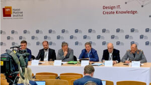 Panel von sechs personen in förmlicher Kleidung, dahinter eine mit HPI-Logos versehene Wand, im Vordergrund Stuhlreihen.