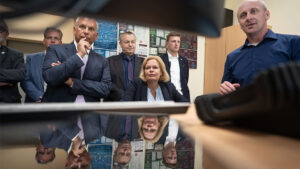 Blick unter einem Monitor hundurch über eine Tischplatte auf Bundesinnenministerin nancy Faeser, umstanden von mehreren Männern in Jackets.