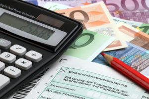 Einkommenssteuerformular, ein Taschenrechner, ein roter Stift und im Hintergrund Geldscheine