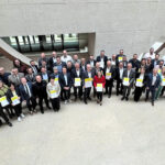 Foto einer größeren menschengruppe in einem Innenraum, alle sind formell gekleidet und halten Dokumente in der Hand.