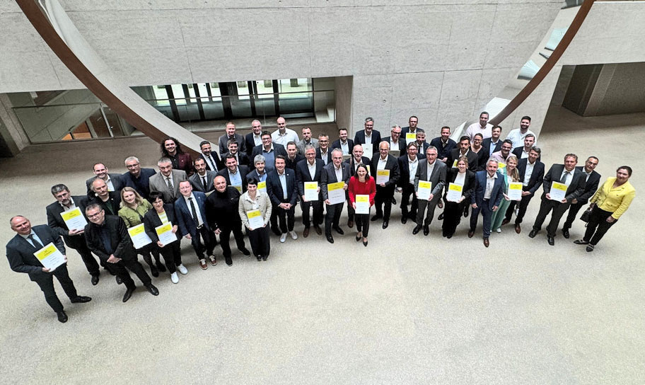 Foto einer größeren menschengruppe in einem Innenraum, alle sind formell gekleidet und halten Dokumente in der Hand.