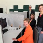 Vier sichtlich gut gelaunte Frauen haben sich in einem nüchternen, grauen Raum mit PC-Cubicles für ein Foto aufgestellt.