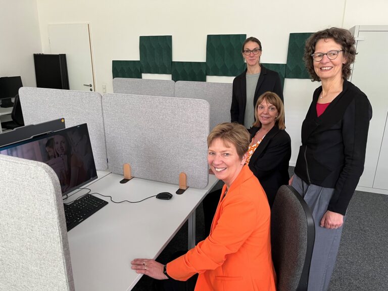 Vier sichtlich gut gelaunte Frauen haben sich in einem nüchternen, grauen Raum mit PC-Cubicles für ein Foto aufgestellt.