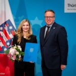 Eine Frau und ein Mann stehen in Business-Kleidung vor einer drapierten Thüringen-Flagge und lächeln in die Kamera.