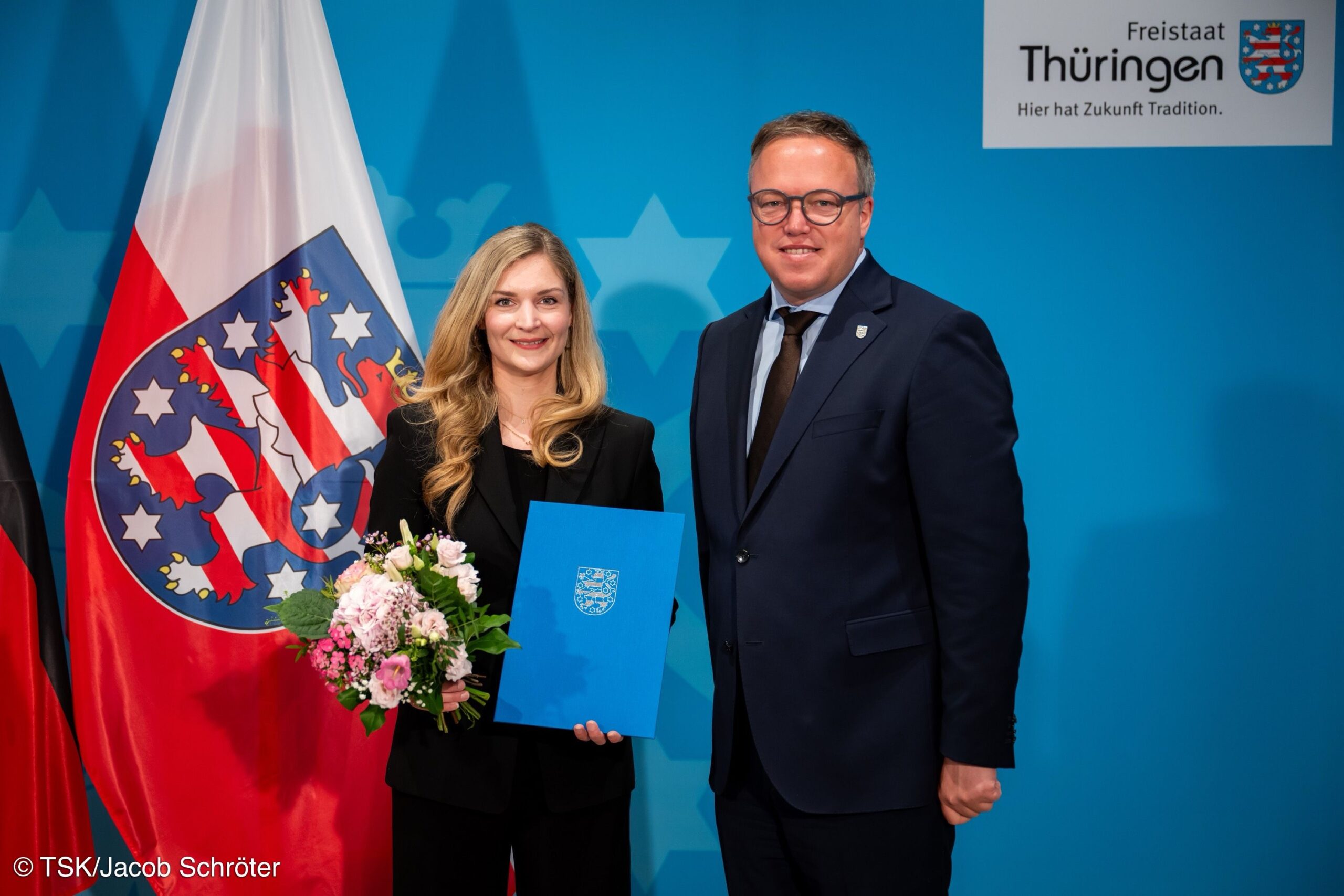 Eine Frau und ein Mann stehen in Business-Kleidung vor einer drapierten Thüringen-Flagge und lächeln in die Kamera.