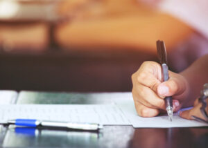 Nahaufnahme einer schreibenden Hand mit Papierstapeln und mehreren Stiften.