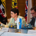 Marion Gentges und Georg Eisenreich sitzen an einem Konferenztisch, im Hintergrund verschiedene Landesflaggen.