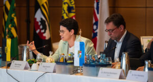 Marion Gentges und Georg Eisenreich sitzen an einem Konferenztisch, im Hintergrund verschiedene Landesflaggen.