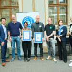 Eine Gruppe von sieben Personen hat sich vor einem Roll-Out-Banner mit dem Umweltzeichen "Blauer Engel" für ein Gruppenfoto aufgestellt. Zwei davon präsentieren Urkunden.