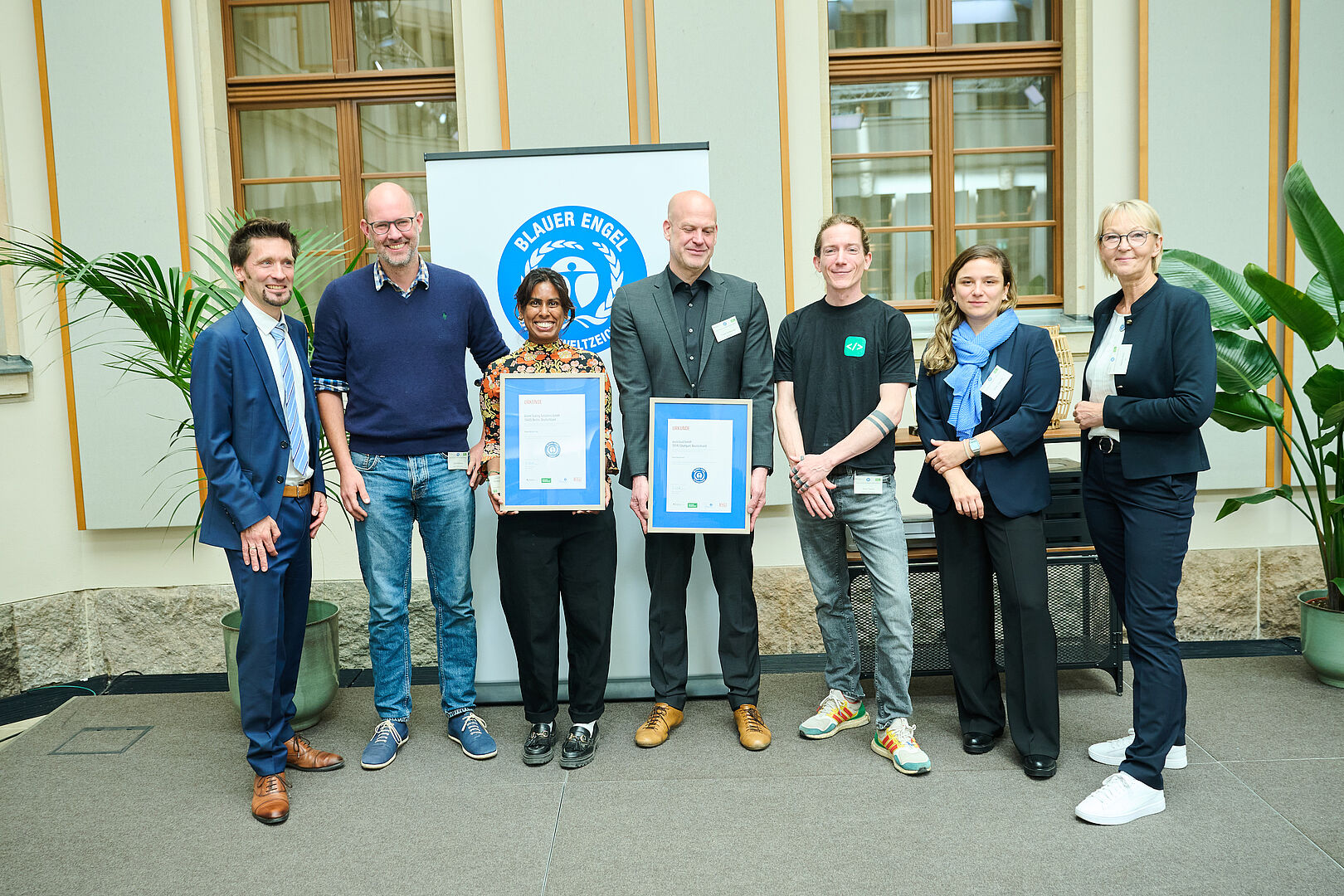 Eine Gruppe von sieben Personen hat sich vor einem Roll-Out-Banner mit dem Umweltzeichen "Blauer Engel" für ein Gruppenfoto aufgestellt. Zwei davon präsentieren Urkunden.