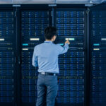Rückenansicht eines Mannes vor einem Serverrack, die Szenerie ist in blaues Licht getaucht.