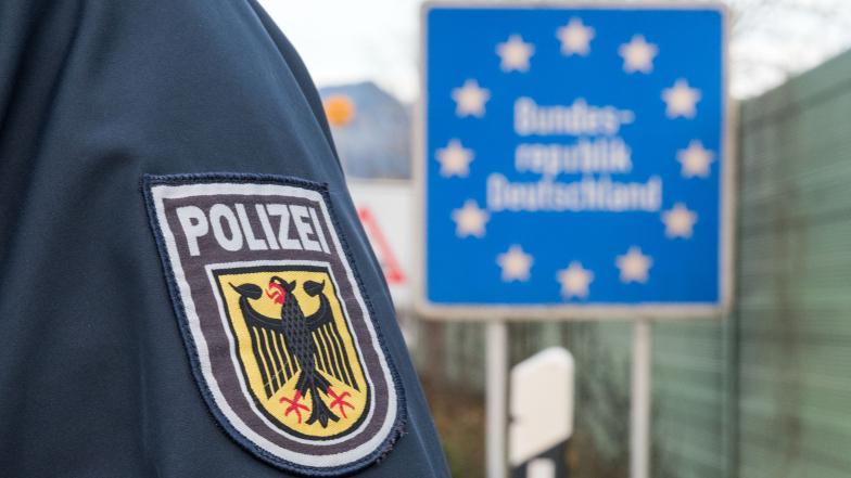 Wappen mit Aufschrift „Polizei“ auf Uniformärmel genäht. Im Hintergrund ein Schild „Bundesrepublik Deutschland“.