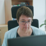 Blick über zwei Monitore auf eine Frau, die in einer Büroumgebung sitzt und anscheinend an ihrem Computer arbeitet.