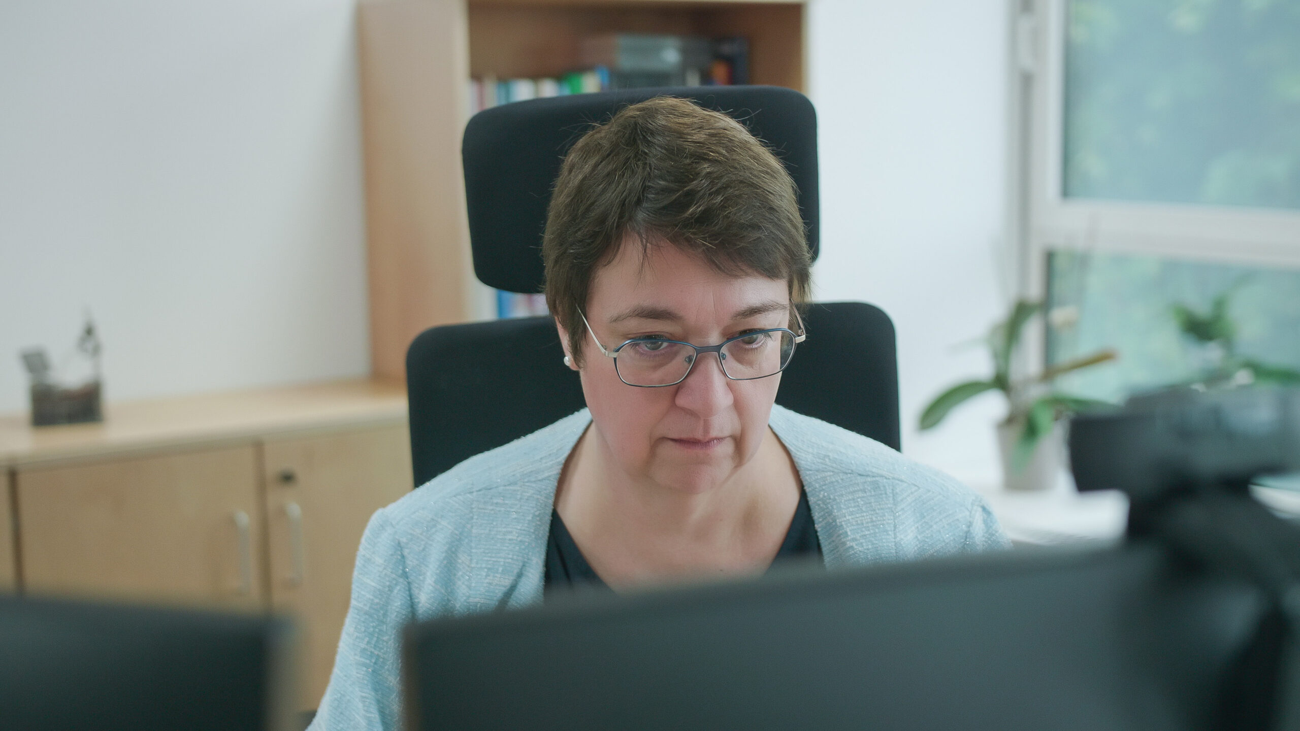 Blick über zwei Monitore auf eine Frau, die in einer Büroumgebung sitzt und anscheinend an ihrem Computer arbeitet.