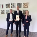 Drei Personen in dunkler, förmlicher Kleidung stehen nebeneinander, der Mann in der Mitte präsentiert eine Urkunde.