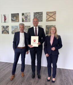 Drei Personen in dunkler, förmlicher Kleidung stehen nebeneinander, der Mann in der Mitte präsentiert eine Urkunde.