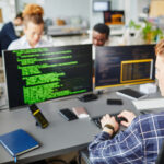 Rückansicht eines jungen, code-schreibenden Mannes und seines Bildschirms, im Hintergrund weitere Personen an Monitoren.
