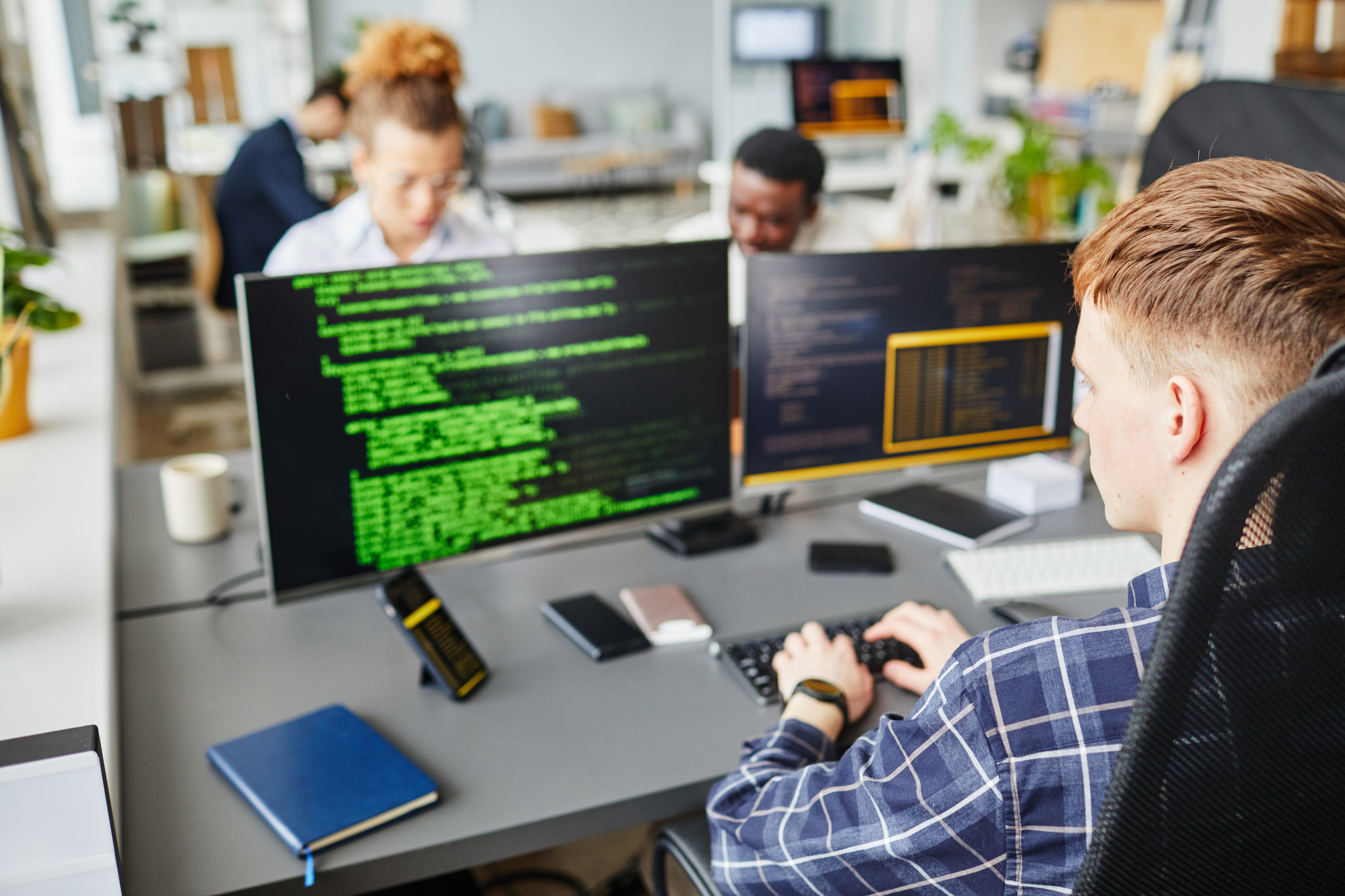 Rückansicht eines jungen, code-schreibenden Mannes und seines Bildschirms, im Hintergrund weitere Personen an Monitoren.
