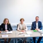 Drei Personen sitzen in einem Raum mit Amtsstuben-Flair nebeneinander an einem Tisch, vor sich Dokumente ausgebreitet.