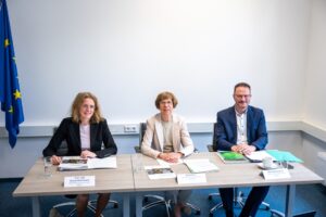 Drei Personen sitzen in einem Raum mit Amtsstuben-Flair nebeneinander an einem Tisch, vor sich Dokumente ausgebreitet.