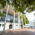 Haupteingang des Bundesrechnungshof-Gebäudes in Bonn. Der weiße, moderne Bau steht unter Bäumen, schlanke Säulen stützen das Vordach.
