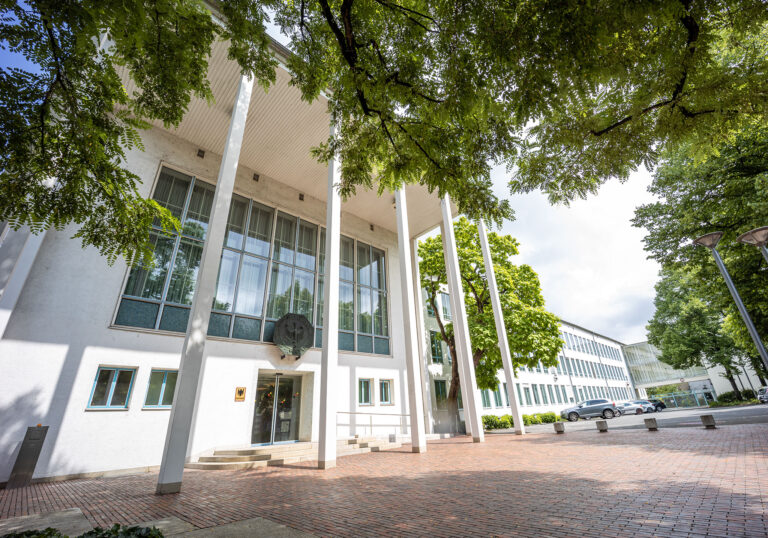 Haupteingang des Bundesrechnungshof-Gebäudes in Bonn. Der weiße, moderne Bau steht unter Bäumen, schlanke Säulen stützen das Vordach.