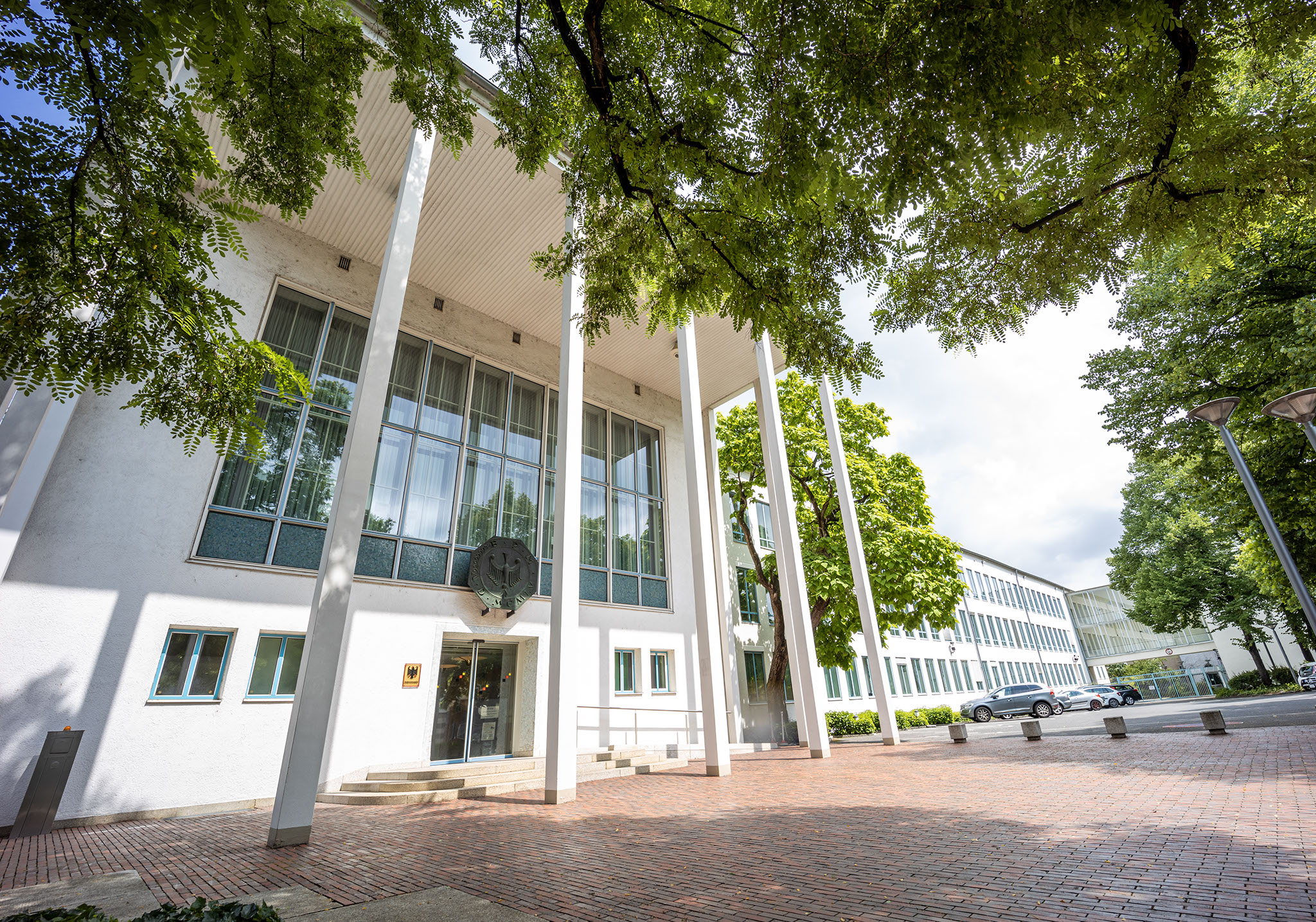 Haupteingang des Bundesrechnungshof-Gebäudes in Bonn. Der weiße, moderne Bau steht unter Bäumen, schlanke Säulen stützen das Vordach.