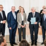 Personen auf einer Bühne, im Vordergrund sieht man Sitzreihen mit Publikum.