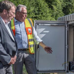 Ein Mann mit Anzug und ein Mann mit Warnweste stehen vor einem geöffneten Elektronik-Schaltkasten und unterhalten sich. Im Hintergrund Himmel und Bäume.