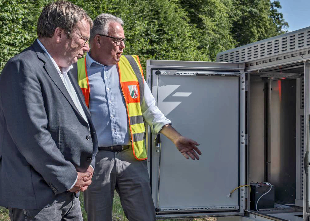 Ein Mann mit Anzug und ein Mann mit Warnweste stehen vor einem geöffneten Elektronik-Schaltkasten und unterhalten sich. Im Hintergrund Himmel und Bäume.