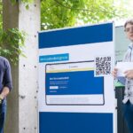 Unter Bäumen stehen zwei junge Männer mit einer großen Präsentationstafel, auf der ein Screenshot der neuen Service-Standard-Website gezeigt wird.
